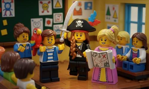 Lila, 6 ans, au sourire fier et tresse châtaine, porte un costume de pirate noir à franges, chapeau à plumes rouges et bleues et brandit une épée scintillante au centre d'une petite scène d'école en bois ; Tom, ~6 ans, cheveux courts bruns, à gauche, en gilet rayé avec un perroquet en plastique sur l'épaule, applaudit ; Anna, ~6 ans, cheveux blonds en couettes, à droite, en robe de princesse, tient une carte au trésor ; Mme Sophie, enseignante dans la trentaine, cheveux attachés, se tient en arrière-plan à gauche devant un tableau, mains jointes ; la salle est lumineuse, décorée de guirlandes et de dessins d'enfants, des parents en briques LEGO sont assis sur des bancs, l'ambiance est festive et inclusive avec un projecteur chaleureux sur la répétition de la pièce.